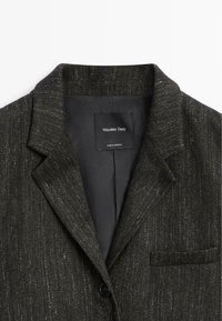 Blazer noir en tissu texturé avec des fils métalliques subtils. Revers cranté, fermeture à un bouton, et poche poitrine.
