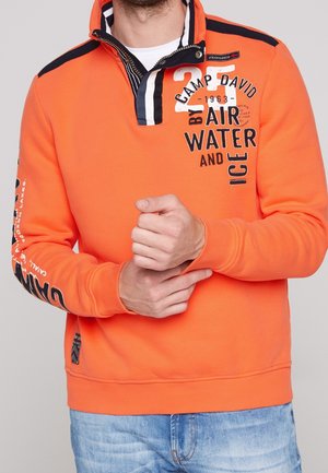 Mężczyzna w pomarańczowym swetrze z czarno-białymi paskami na kołnierzu oraz białym, czarnym napisem "Camp David Air Water and Ice" na piersi i rękawie.