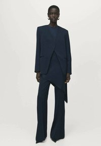 Tummansininen räätälöity puku, jossa on jämäkkä bleiseri terävillä läpillä ja leveät housut, valmistettu tekstuurista kankaasta, jossa on sileä pinta.