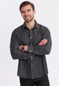 Chemise boutonnée gris charbon en tissu texturé, avec des boutons blancs, une poche poitrine et des manches longues, accompagnée d'une montre noire.
