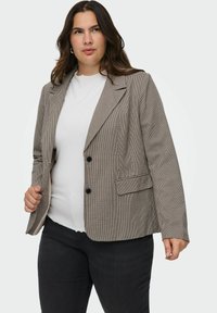 Žena v béžovém kostkovaném saku přes bílý žebrovaný top a černé kalhoty, stojící proti jednoduchému světlému pozadí.