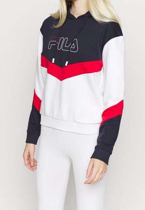Person iført en sort, rød og hvid FILA hættetrøje med snore, kombineret med hvide leggings, vist fra skuldre til hofter.