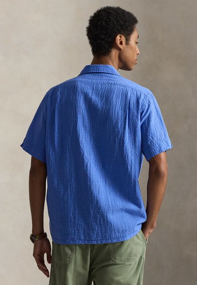 Homme aux cheveux bouclés portant une chemise bleue texturée à manches courtes et un pantalon vert, debout les mains près des poches, de dos.