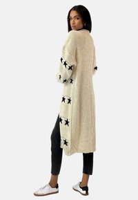 Cárdigan de punto beige con abertura frontal, que presenta patrones de estrellas negras en las mangas. Combinado con jeans oscuros y zapatillas blancas.