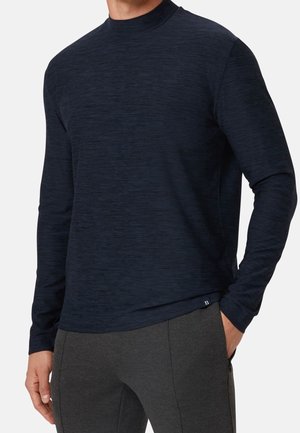 Homme portant une chemise moulante à manches longues et col montant bleu marine foncé, ainsi qu'un pantalon gris, avec une main dans la poche.