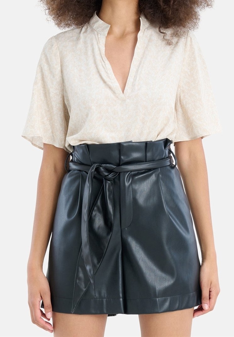 Světlé béžové blůzka s krátkým rukávem a výstřihem do V, se slabým vzorem, zkombinovaná s tmavozelenými šortkami z umělé kůže s vysokým pasem a zauzlovaným opaskem.