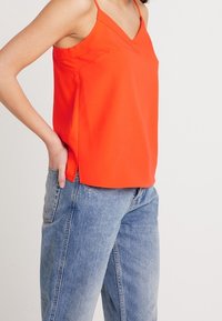 Haut sans manches orange vif avec col en V, fentes latérales et texture lisse, associé à un jean en denim bleu clair.