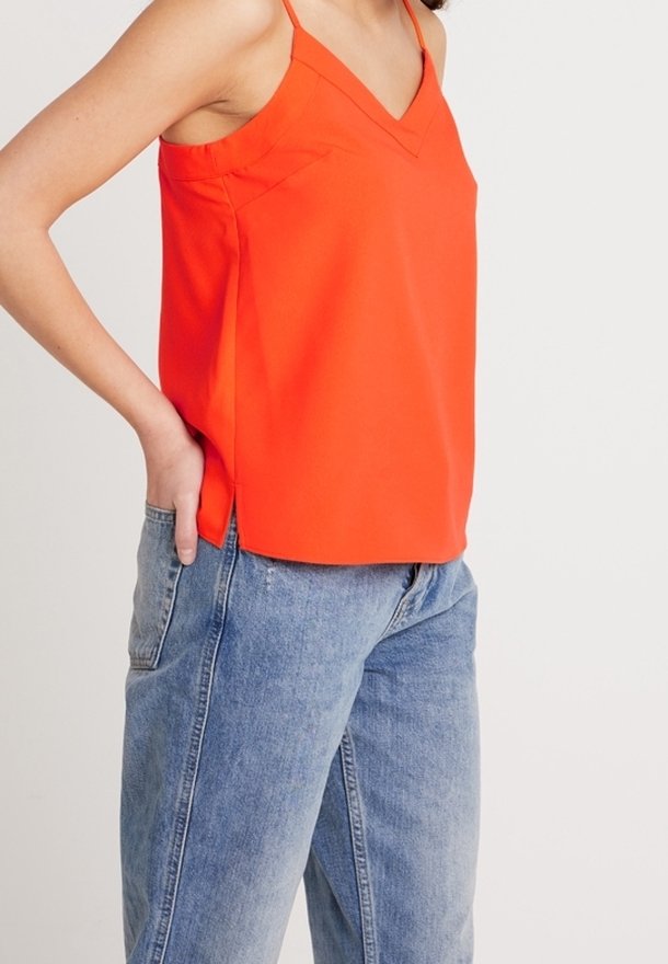 Haut sans manches orange vif avec col en V, fentes latérales et texture lisse, associé à un jean en denim bleu clair.