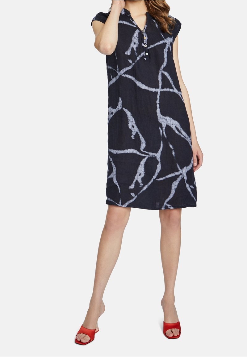 Femme portant une robe noire mi-longue avec des motifs abstraits blancs et des boutons au niveau de l'encolure, associée à des sandales rouges à talons ouverts.