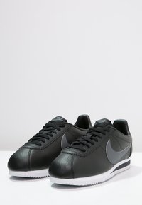 Baskets en cuir noir avec une finition texturée, accent du logo Nike gris, lacets noirs et semelle en caoutchouc blanche avec motif de crampons en zigzag.