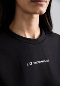 Μαύρο βαμβακερό μπλουζάκι με στρογγυλή λαιμόκοψη και λευκή εκτύπωση λογότυπου: "EA7 EMPORIO ARMANI" στο στήθος. Απαλή υφή.