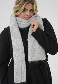 Sciarpa a maglia di un grigio chiaro e texture morbida, appoggiata attorno al collo, abbinata a un cappotto nero con bottoni.