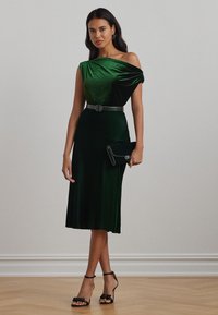 Green velvet dress with one off-shoulder sleeve, fitted bodice, flared skirt, and a black belt; paired with a matching clutch and black heels.