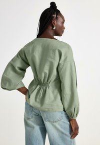 Lys grøn, langærmet bluse med samlet talje. Blødt stof, løs pasform og ballonærmer. Båret med slidte blå jeans.