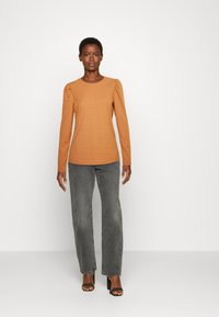 Top de manga longa laranja queimado com textura canelada e ombros bufantes, combinado com jeans retos cinza de cintura alta e saltos pretos.