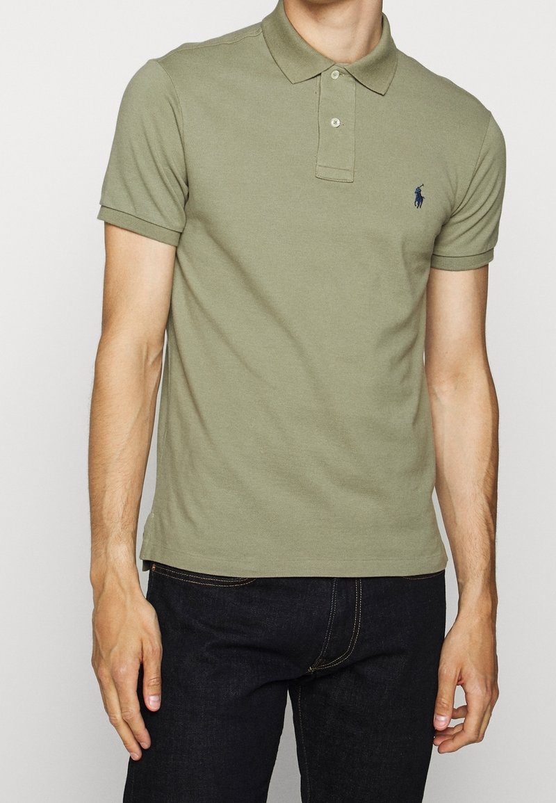 Homme portant un polo vert olive à manches courtes avec un petit logo brodé foncé sur la poitrine gauche.
