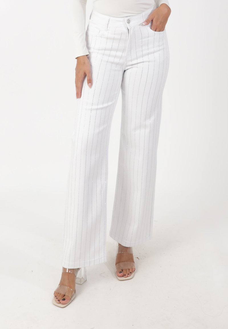 Pantalones blancos de pierna ancha con rayas verticales plateadas, hechos de una tela suave. Presentan bolsillos delanteros y cintura de talle medio. Sandalias de tacón transparentes.