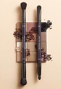 Zwei schwarzgriffige Lidschattenpinsel mit weichen Borsten, die über einer Palette von neutralen und schimmernden Farbtönen mit verstreuten Produkten positioniert sind.