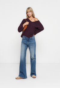 Pull violet à épaules dénudées avec texture côtelée, associé à un jean flare bleu clair et des mocassins beiges. Évoque un look décontracté.