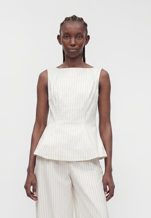 Femme aux cheveux tressés portant un top blanc sans manches à fines rayures avec basque et un pantalon large assorti, debout devant un fond uni.