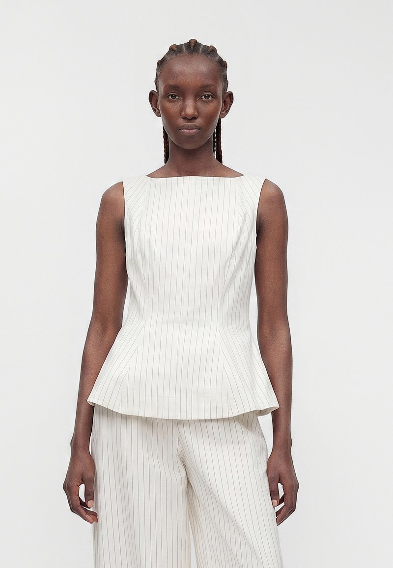 Femme aux cheveux tressés portant un top blanc sans manches à fines rayures avec basque et un pantalon large assorti, debout devant un fond uni.