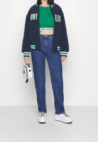 Námořnický fleecový bomber, zelené zkrácené tričko, modré džíny s vysokým pasem, bílé tenisky, držící malou bílou tašku.