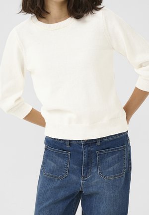 Femme portant un pull blanc en maille à manches longues et un jean en denim bleu avec des poches avant, debout, les mains derrière le dos.