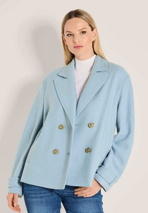 Frau in einem hellblauen doppelreihigen Wollmantel mit goldenen Knöpfen und einem weißen Rollkragenpullover, posiert mit der Hand auf der Hüfte.