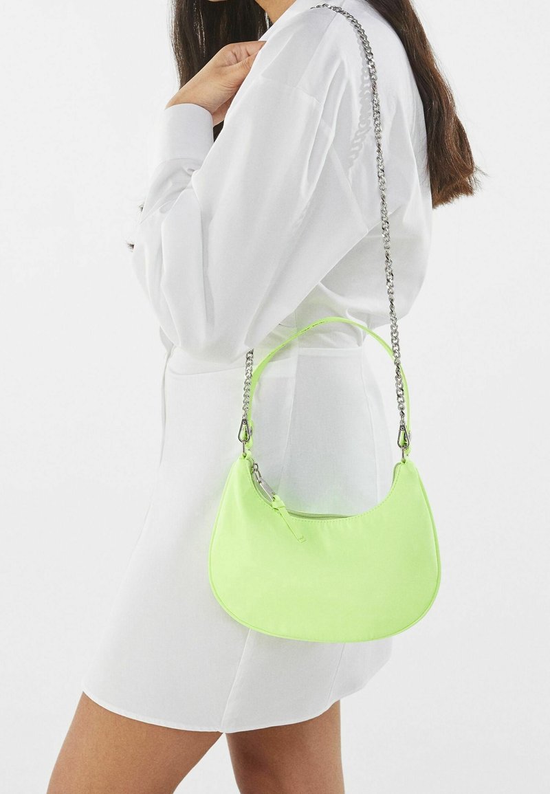 Femme en robe blanche portant un petit sac à main vert fluo avec une anse chaîne argentée sur l'épaule.