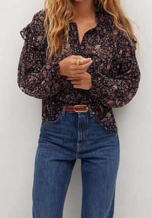 Femme portant un jean bleu taille haute, une ceinture marron et un chemisier sombre à manches longues à motif floral avec des épaules à volants, mains jointes à la taille.