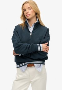 Sweat-shirt à col zippé quart bleu marine avec des accents côtelés. Doté d'un logo brodé, d'une texture douce, superposé à une chemise à rayures bleu clair.