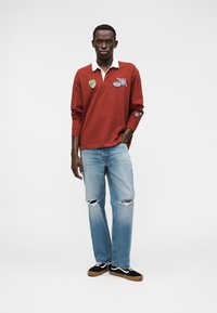 Κόκκινο μακρυμάνικο πόλο με λευκό γιακά και μπαλώματα, συνοδευόμενο από ανοιχτό μπλε τζιν με σκισίματα και μαύρα αθλητικά παπούτσια με λευκές λεπτομέρειες.