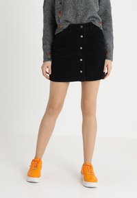 Personne portant un pull gris avec des détails arc-en-ciel, une jupe en velours côtelé noire à boutons et des baskets orange vif, se tenant devant un fond blanc.