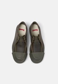 Scarpe di gomma marroni con punta arrotondata, contrasti di copertura in gomma scura e solette beige con testo rosso "CAMPE". Cuciture decorative visibili.