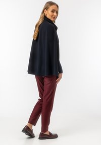 Granatowy wełniany płaszcz z wysokim kołnierzem, połączony z dopasowanymi burgundowymi spodniami i ciemnymi butami typu slip-on, prezentując gładką, teksturowaną tkaninę.