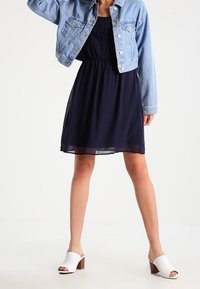 Marinblå klänning med samlad midja, tillsammans med en ljusblå denimjacka. Vita öppna-toe mule-skor med en brun klossig klack.