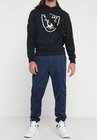 Felpa nera con logo bianco dei Raiders, abbinata a pantaloni della tuta blu navy. Sneakers bianche con dettagli neri completano il look. Borsa a tracolla regolabile.