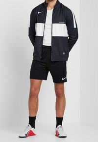 Zwarte Nike windbreaker met witte horizontale strepen, gecombineerd met zwarte shorts. Sport schoenen met rode accenten. Eenvoudig wit t-shirt eronder.
