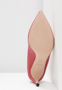 Chaussure à talon haut en cuir rouge avec un bout pointu et une semelle beige, dotée d'un petit grip noir au bas du talon.