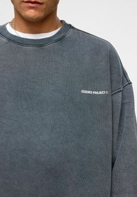 Grijze katoenen sweatshirt met verlaagde schouders, geribde halslijn en de tekst "ESSENCE PROJECT" op de borst in het wit. Zachte textuur, casual pasvorm.