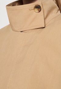 Col montant beige attaché avec un gros bouton en écaille, montrant des coutures détaillées sur un tissu lisse.