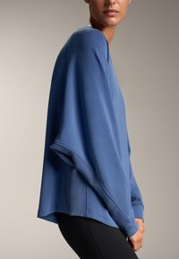 Modra velika majica s kapuco z ribanimi manšetami in stranskimi poudarki. Mehka tkanina, kvadratna silhueta in spuščena ramena ustvarjajo sproščen videz.