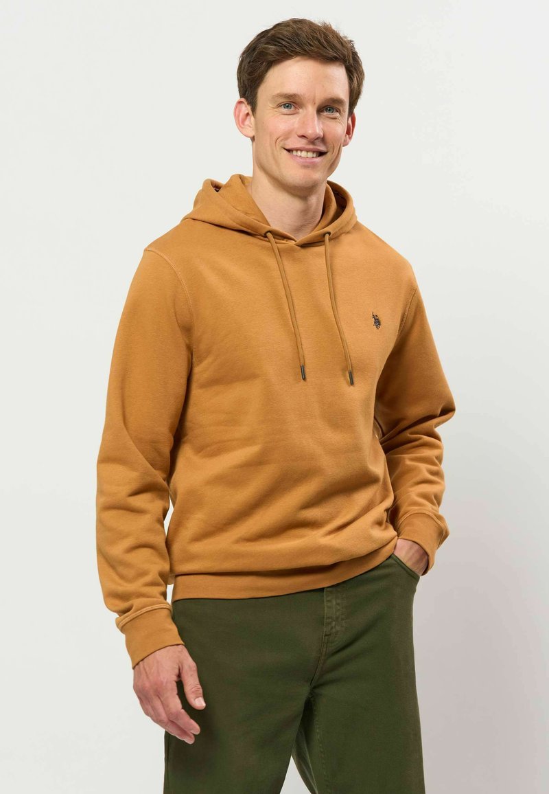 Jeune homme souriant, portant un sweat à capuche marron et un pantalon vert, debout avec une main dans la poche devant un fond uni.