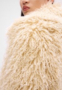 Textuurrijke, crèmekleurige faux fur jas met een volumineus, krullend ontwerp, dat ingewikkelde details in de stof laat zien.