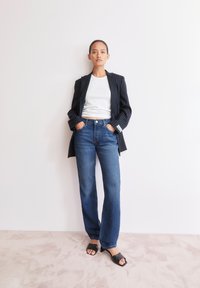 Blazer noir à rayures sur un crop top blanc, associé à un pantalon large en denim bleu et des sandales à talons noires, sur un fond clair.