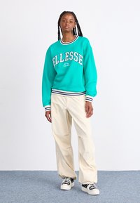 Τιρκουάζ φούτερ με το λογότυπο "Ellesse", ριγέ ribbed μανίκια και λαιμόκοψη, σε συνδυασμό με κρεμ παντελόνι φαρδιάς γραμμής και μαύρα-άσπρα αθλητικά παπούτσια.