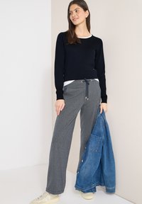 Femme debout contre un mur, portant un pull noir, un pantalon gris à cordon de serrage, des baskets blanches, tenant une veste en jean bleue dans une main.