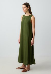 Kobieta stojąca bokiem w bezrękawną, oliwkową sukienkę do kostek i brązowe płaskie sandały na neutralnym, jasnym tle.