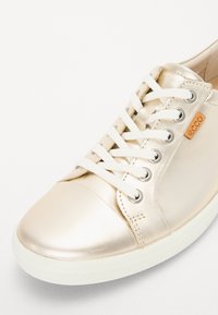 Metallic gold sneaker with white laces, rounded toe, and a rubber sole. Notable details include silver eyelets and a tan brand tag.