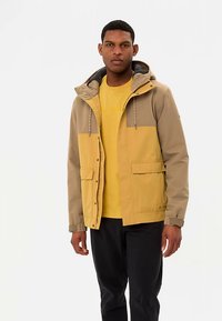 Veste jaune et marron avec capuche, dotée de deux poches avant, de cordons décoratifs et d'une doublure intérieure douce. Portée par-dessus une chemise jaune.